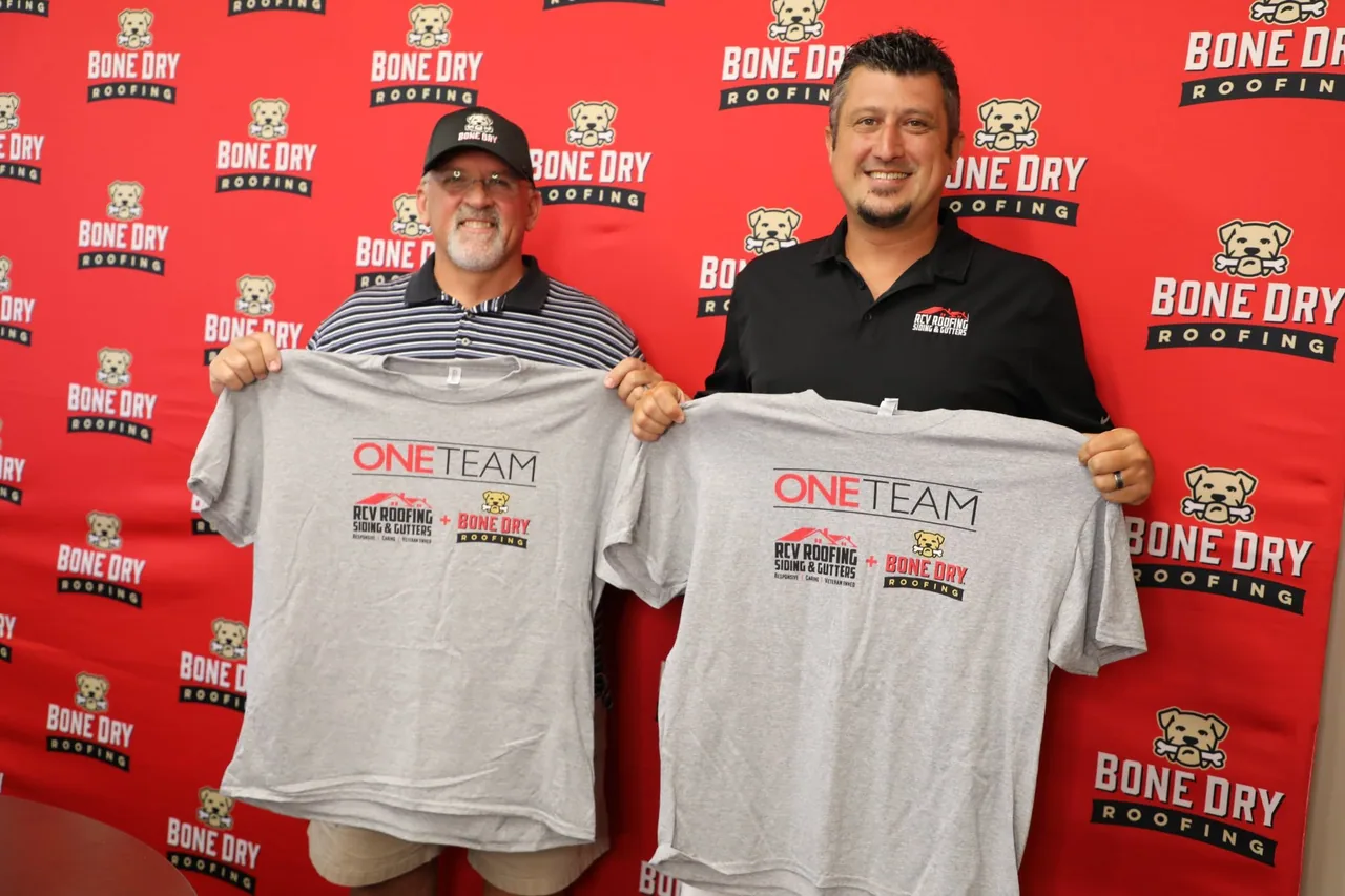 Two people holding One Team shirts for RCV Roofing, Siding & Gutters and Bone Dry Roofing in front of a Bone Dry Roofing backdrop